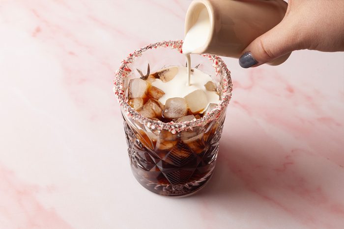 3/4th shot of a hand pours cream into iced coffee and the ice cubes float at the top while the glass shows a red and white sprinkle rim nearby pink marble