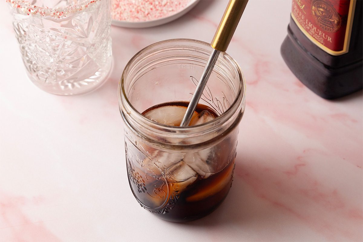 3/4th shot of a mason jar with iced coffee and a metal straw rests on a pink marbled surface Nearby are a glass cup a liqueur bottle and candy pieces