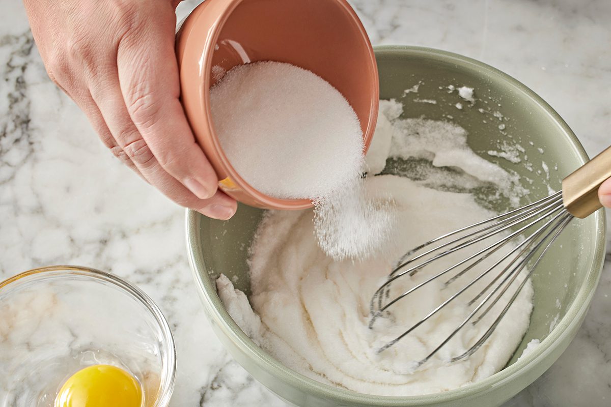 A person pours granulated sugar from a small bowl into a mixing bowl with a creamy mixture, while holding a whisk. An egg sits in a separate glass bowl on a marble countertop.