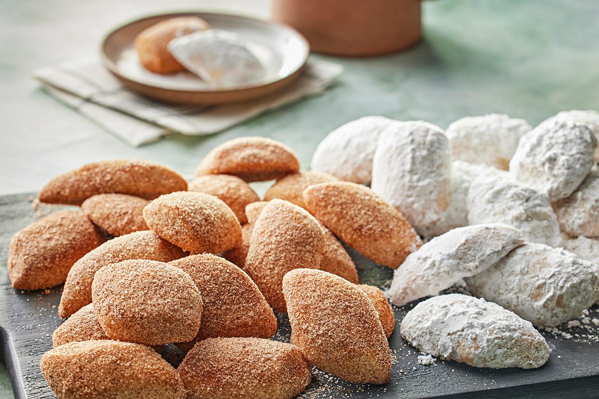 A platter with two types of crescent-shaped cookies: one coated in cinnamon sugar and the other dusted with powdered sugar, with more cookies on a plate in the background.