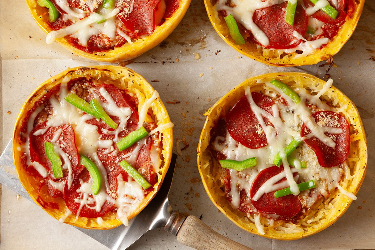 Overhead close-up shot of baked spaghetti squash pizzas topped with melted cheese, pepperoni, and sliced green peppers; A metal spatula slides under one squash half, revealing its golden, textured edges;