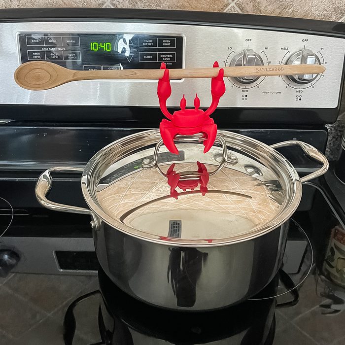 A red crab-shaped utensil holder is clipped to the rim of a stainless steel pot, holding a wooden spoon horizontally above the pot