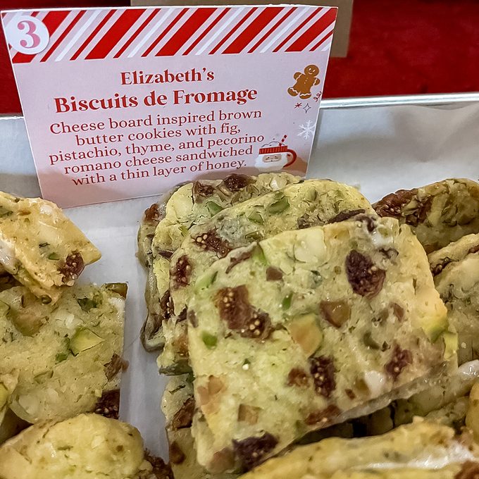 A tray of rectangular, brown butter cookies with visible pieces of fig, pistachio, and herbs. A sign behind them reads "Elizabeth's Biscuits de Fromage" and lists the ingredients and features of the cookies.